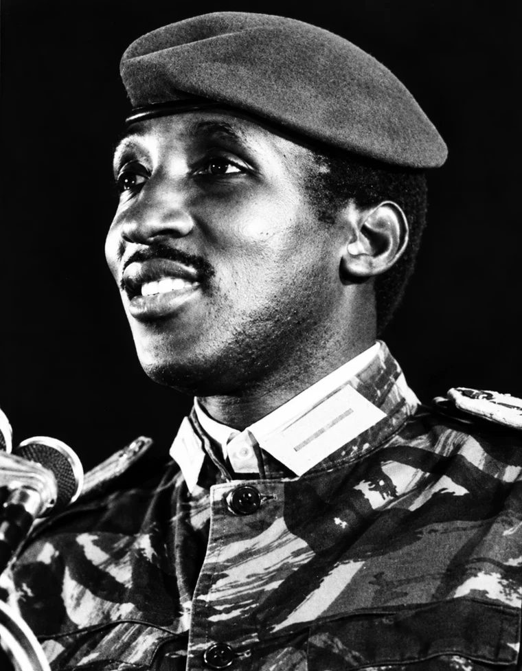 Le Camarade Président Thomas Sankara prenant la parole à Harlem, à New York, en 1984. En treillis de camouflage et béret, il parle au micro, le regard levé, souriant.