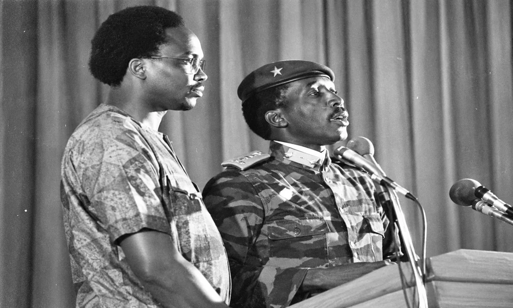 Le Camarade Président Thomas Sankara à une tribune, devant plusieurs micros. Il porte un treillis de camouflage, des pattes d'épaule et un béret à l'étoile. Un homme à lunettes, en chemise à motifs, se tient à sa gauche et traduit.