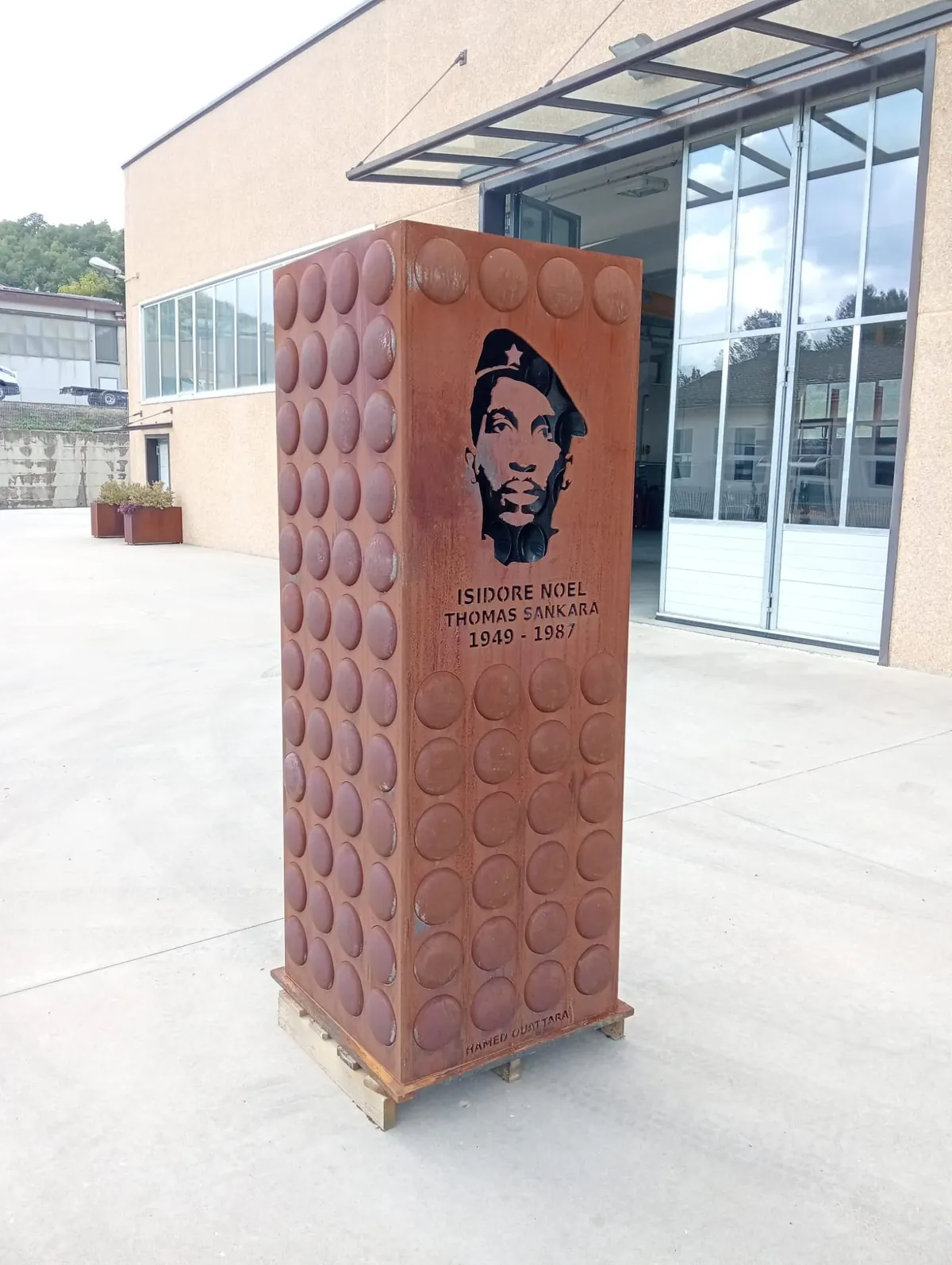 Le monument SANKARA, L'ÉTERNEL VEILLEUR de Hamed Ouattara achevé, en acier Corten oxydé : la face portrait montre le visage du Camarade Président Thomas Sankara découpé au travers de la tôle, au-dessus du bloc portant ISIDORE NOEL THOMAS SANKARA 1949-1987, et la face voisine est couverte de bossages hémisphériques.