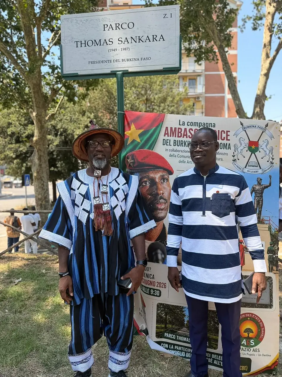 Sous la plaque du Parco Thomas Sankara, Via Ugo della Seta à Rome, deux hommes devant l'affiche de la rencontre préparatoire : le Camarade Madi Sakandé, en tenue traditionnelle burkinabè tissée et coiffe, et le Camarade Yemboini Étienne Lompo. La plaque porte PARCO THOMAS SANKARA 1949-1987 PRESIDENTE DEL BURKINA FASO.